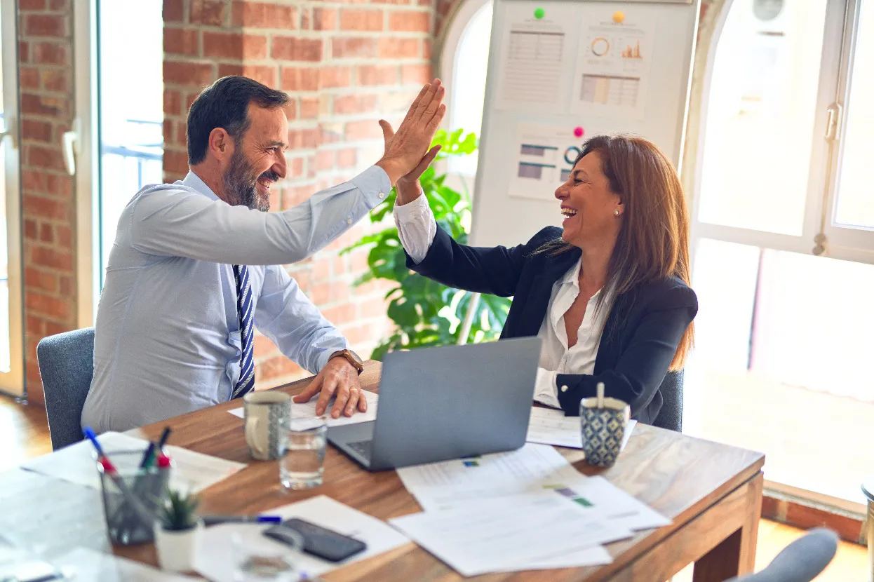 Two business individuals enthusiastically high five at a desk, representing the spirit of teamwork and collaboration in the workplace.