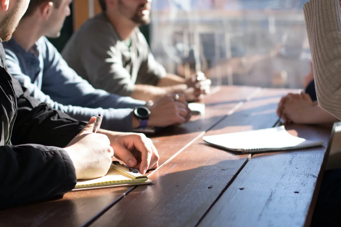 A team of people from various backgrounds gathers at a table, sharing ideas while referencing a notebook