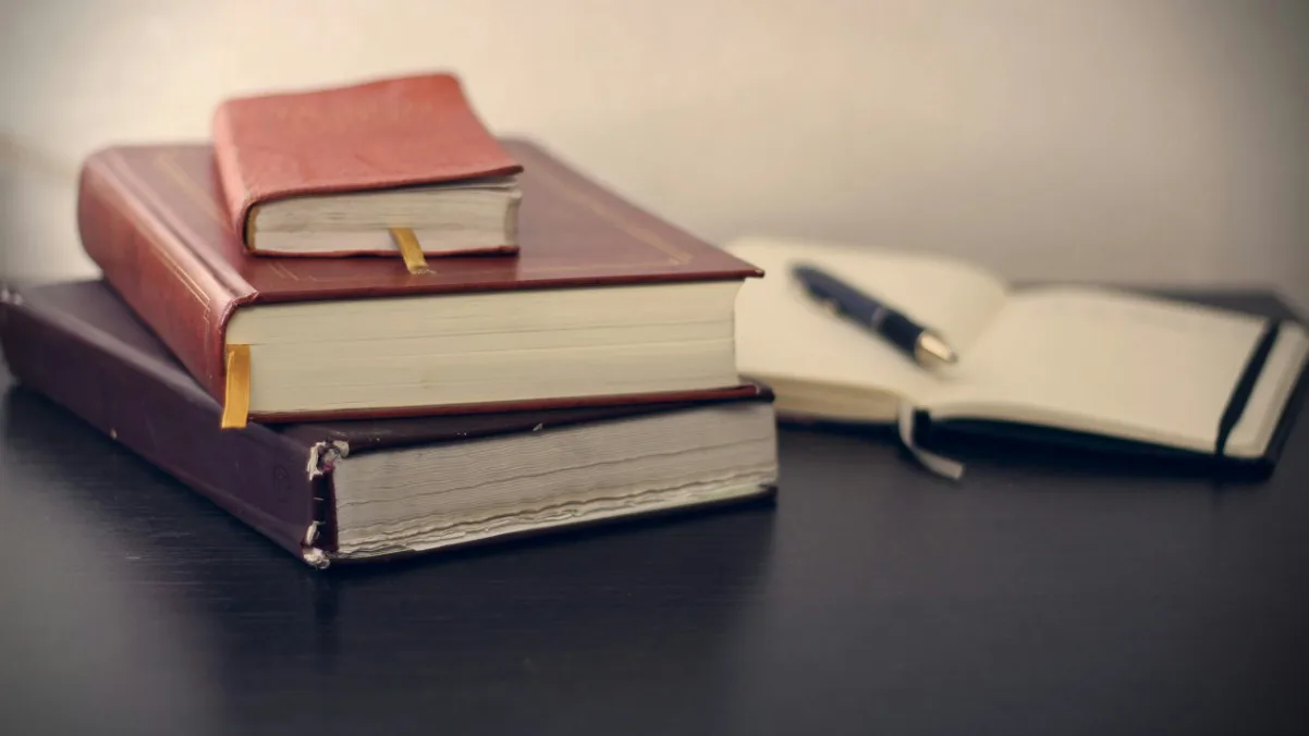 Three books resting on a table alongside a pen and an open notebook